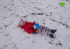 Usmiechnięta Oliwia leży na śniegu po zjeździe na listku z górki saneczkarskiej.